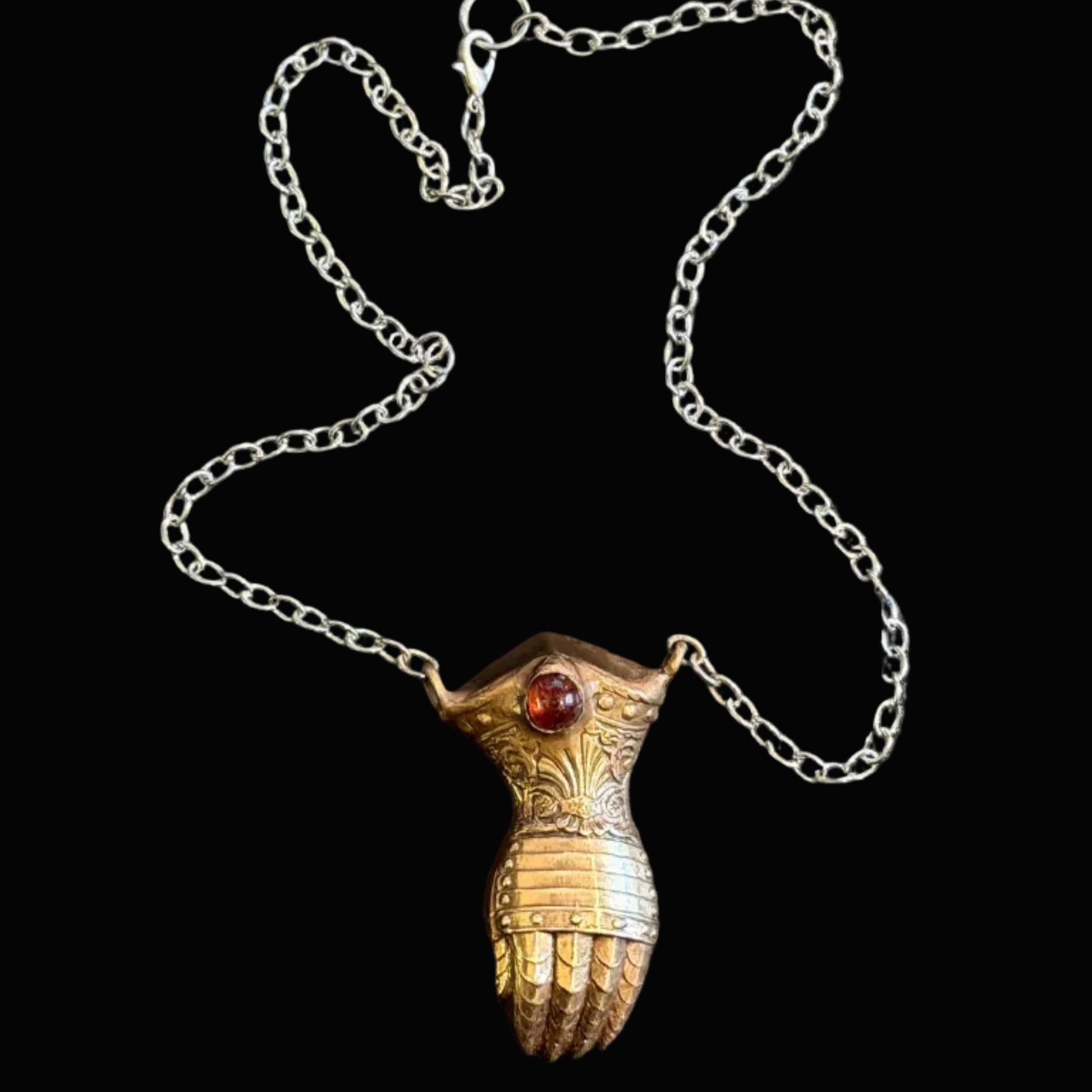 A silver chain necklace featuring a pendant shaped like a stylized hand with a small red gemstone, displayed on a dark textured background.