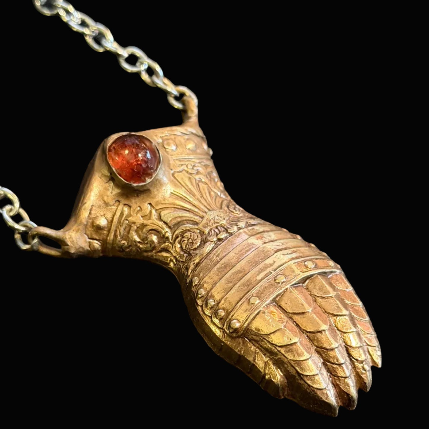 A close-up of a pendant necklace featuring a sculpted owl head in a warm metallic finish, with a small round amber-red gemstone set in its eye socket, hanging from a silver-toned chain against a dark textured background.