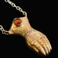 A close-up of a pendant necklace featuring a sculpted owl head in a warm metallic finish, with a small round amber-red gemstone set in its eye socket, hanging from a silver-toned chain against a dark textured background.