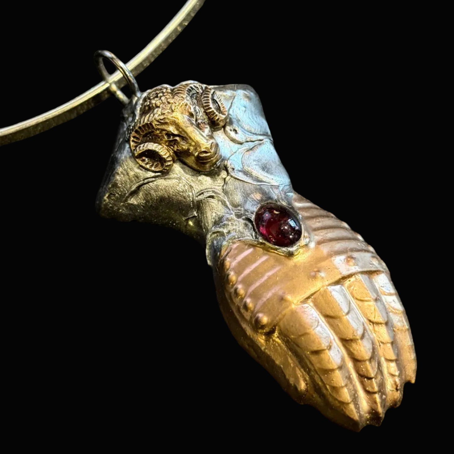 A close-up of a handmade pendant necklace featuring a textured, multi-colored bird or frog figure with a silver-toned metal body, gold-toned wing details, and a small red gemstone, suspended from a thin silver wire on a dark textured background.