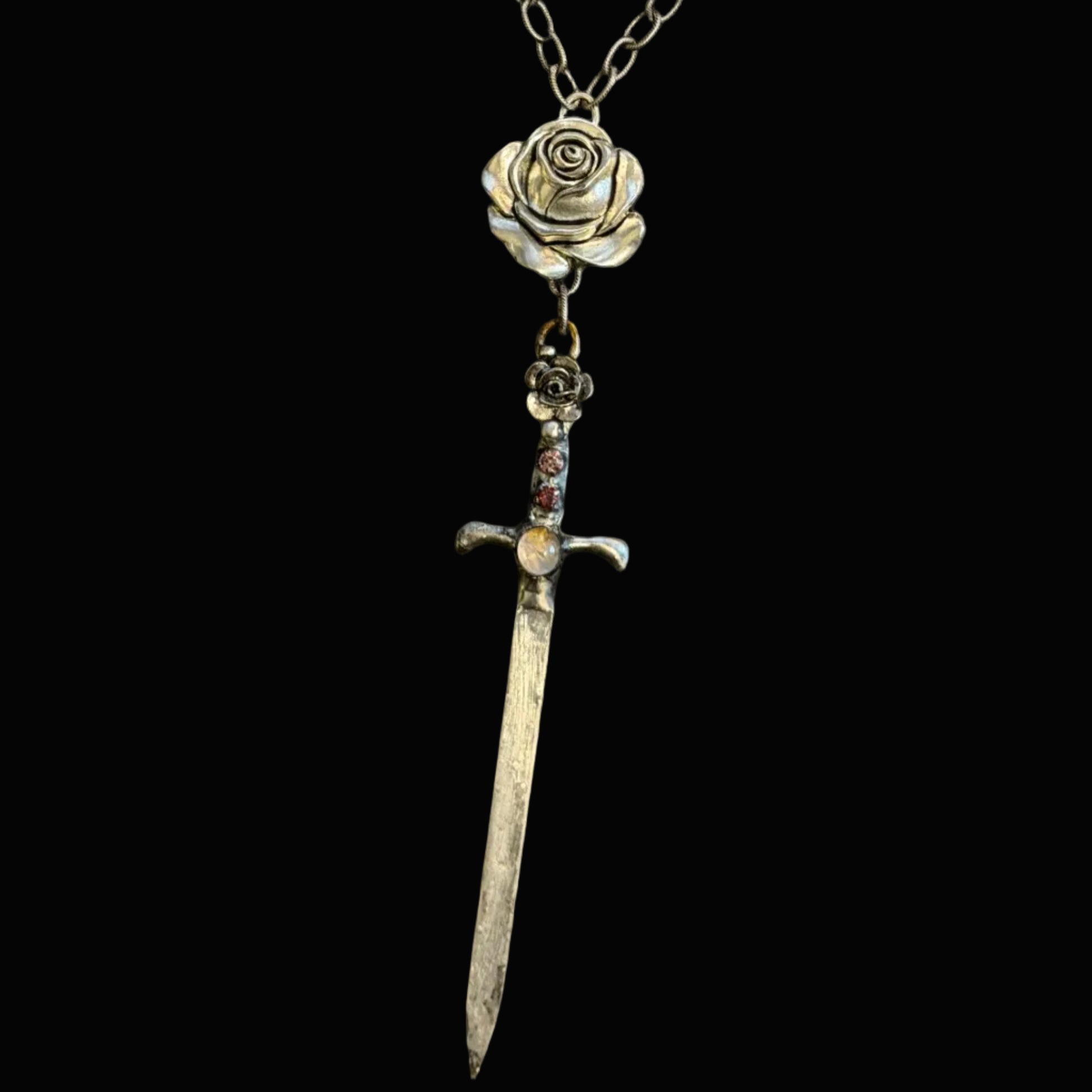 A silver-toned chain necklace featuring a sculpted rose pendant and a dangling sword pendant, displayed against a dark textured background.