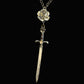 A silver-toned chain necklace featuring a sculpted rose pendant and a dangling sword pendant, displayed against a dark textured background.