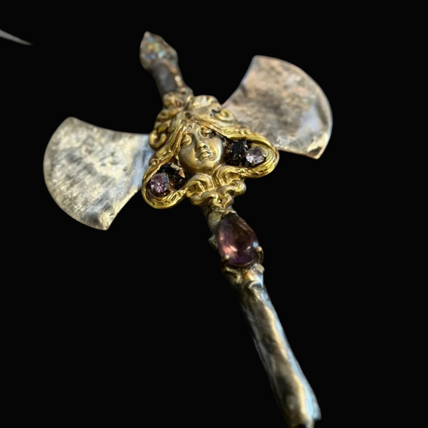 A close-up of a decorative axe pendant necklace with a silver-toned blade, gold-toned hilt, and small purple gemstones, shown on a dark textured background.