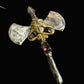 A close-up of a decorative axe pendant necklace with a silver-toned blade, gold-toned hilt, and small purple gemstones, shown on a dark textured background.
