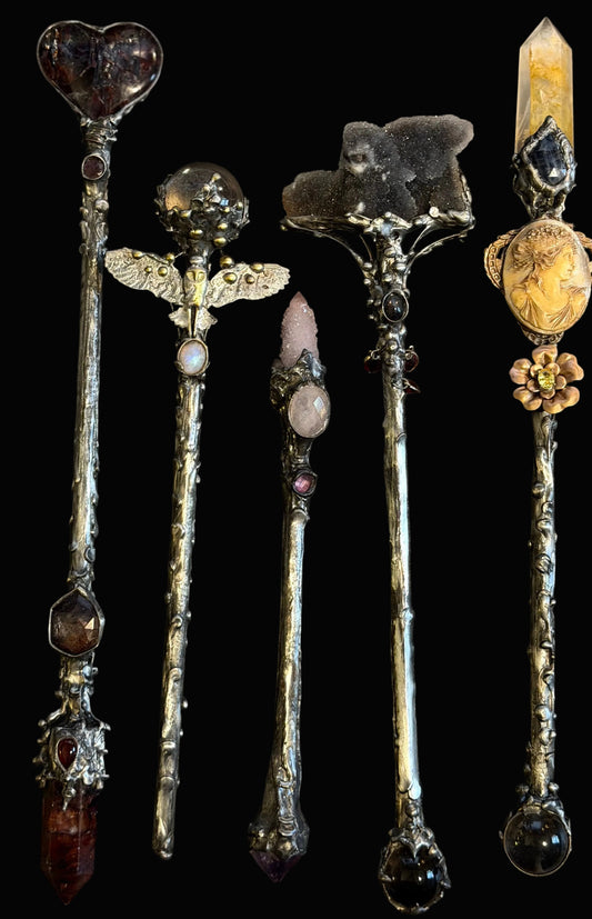 Five ornate silver-toned crystal and gemstone wands arranged vertically on a dark background. The wands feature unique pendants including a heart-shaped stone, angel wings, a pink stone, a dark cat skull, and a yellow stone with floral detailing.