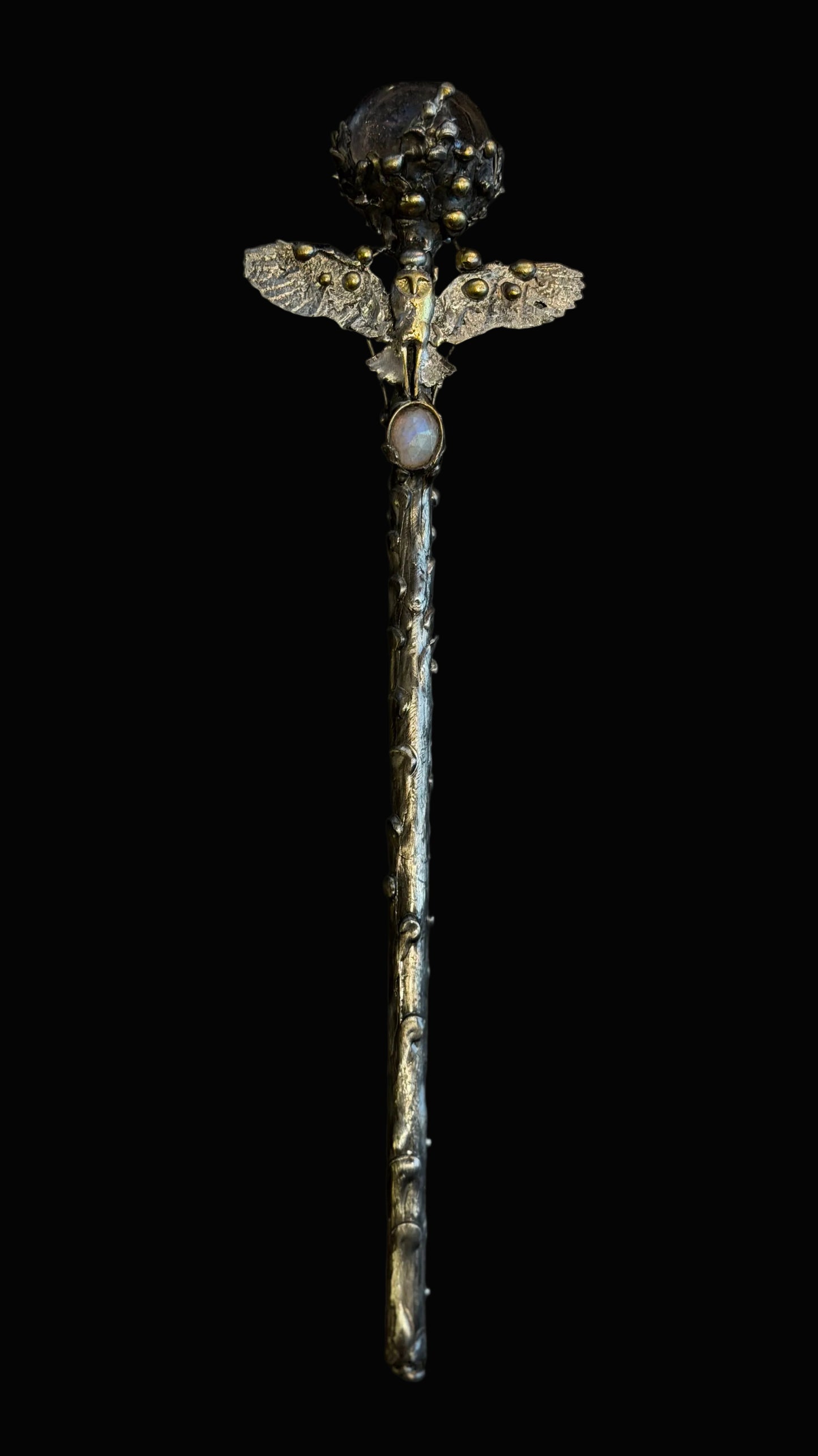 A decorative silver-toned staff or wand with a textured stem, a winged top detail, and a dark gemstone or crystal at the end, photographed against a black background.