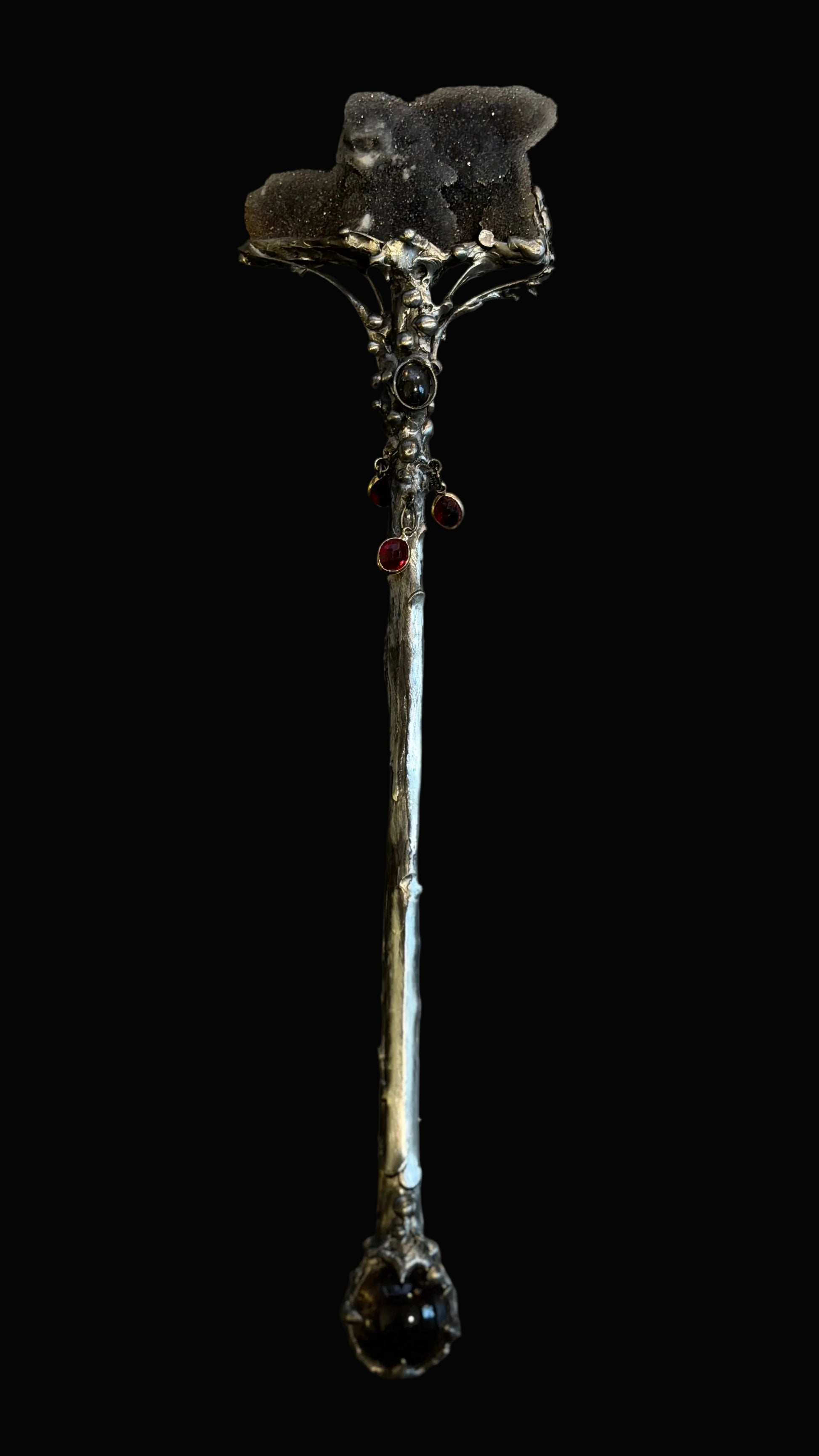 A gothic-style decorative staff or wand with a dark, textured top, small red gem accents, and a faceted dark gemstone at the bottom, photographed against a dark background.