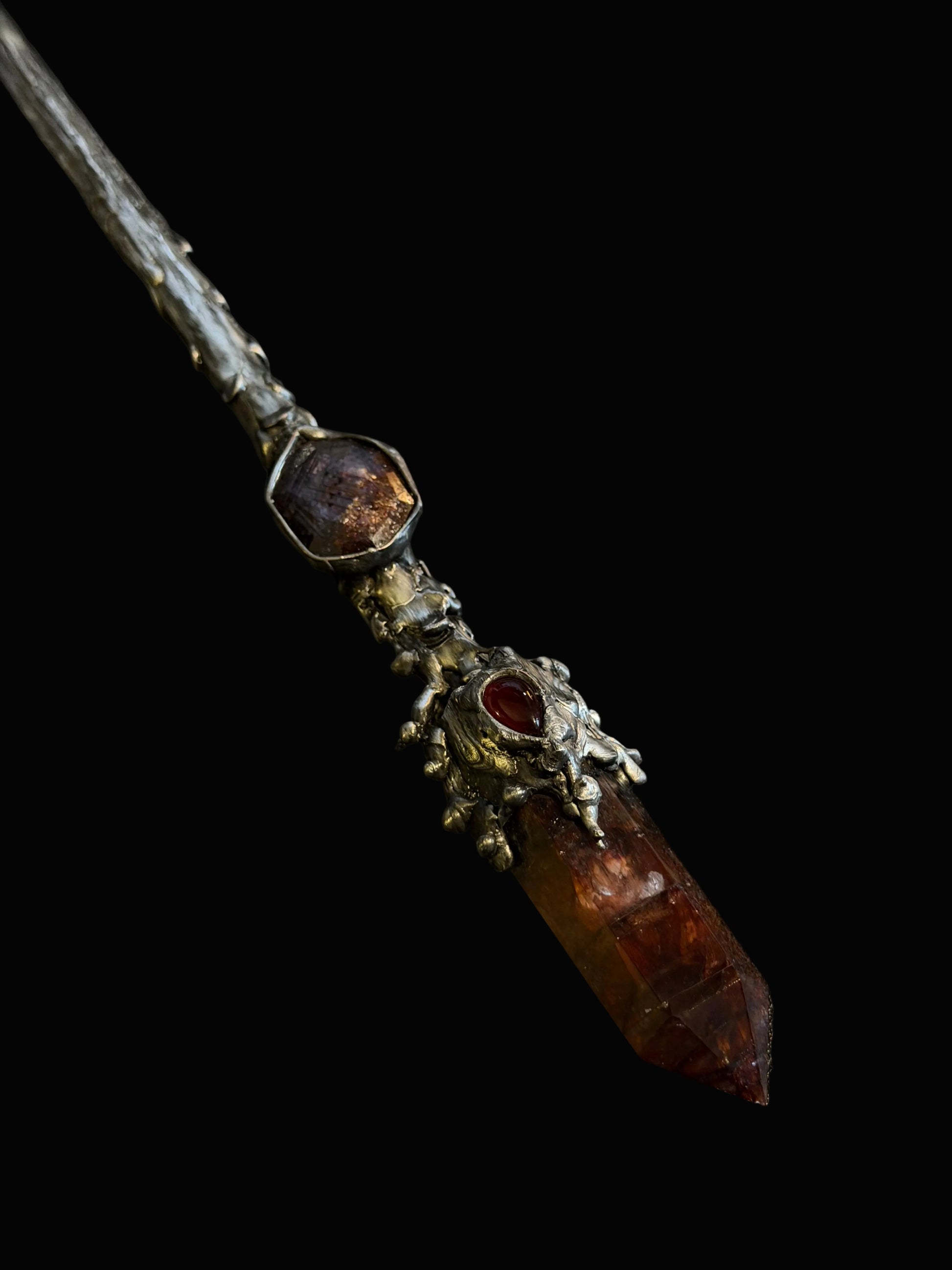 A gothic-style silver wand featuring a large faceted amber crystal point and a smaller reddish gemstone, photographed on a dark textured background.