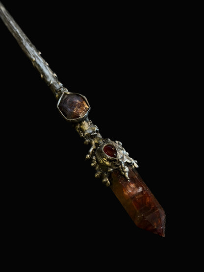 A gothic-style silver wand featuring a large faceted amber crystal point and a smaller reddish gemstone, photographed on a dark textured background.