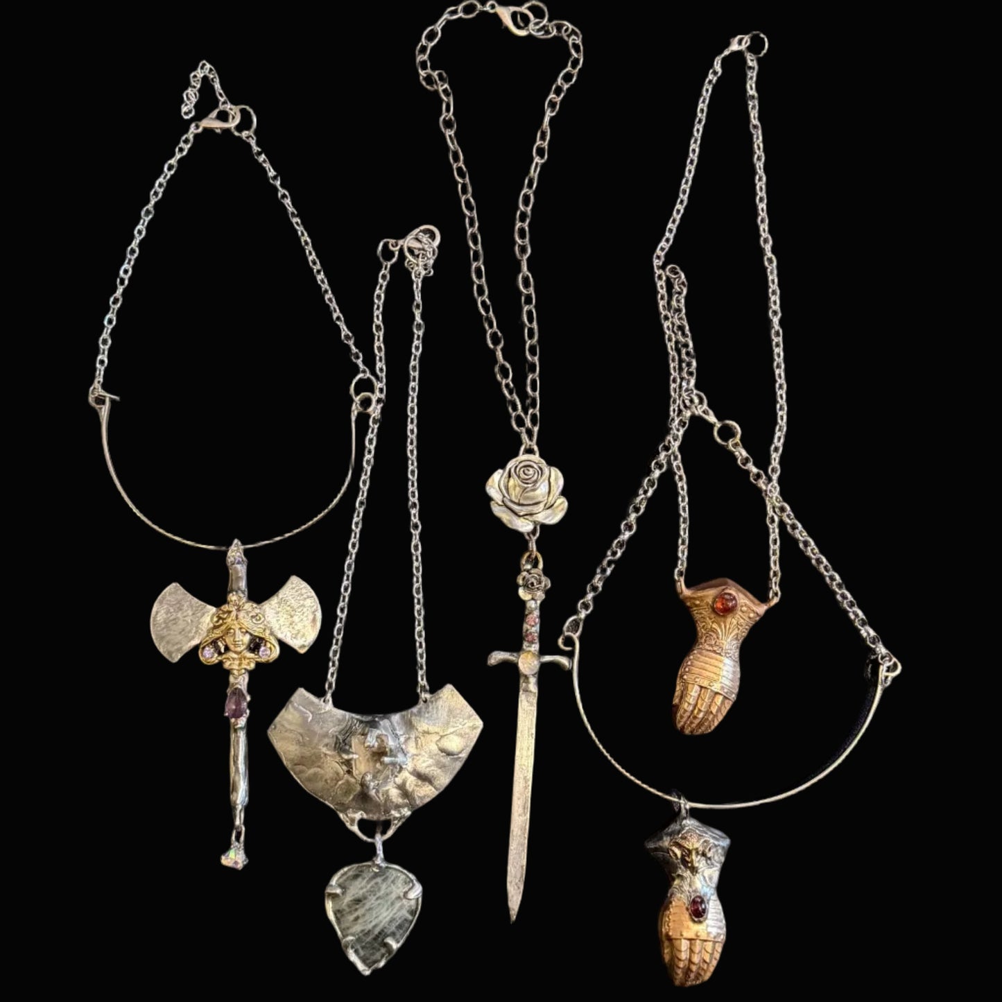 Four distinct silver-toned pendants on chains laid out on a dark desk surface, with a keyboard at the top and dark clothing at the bottom. The pendants include a winged shield, a sword, a rose, a bird, and a small animal figure.