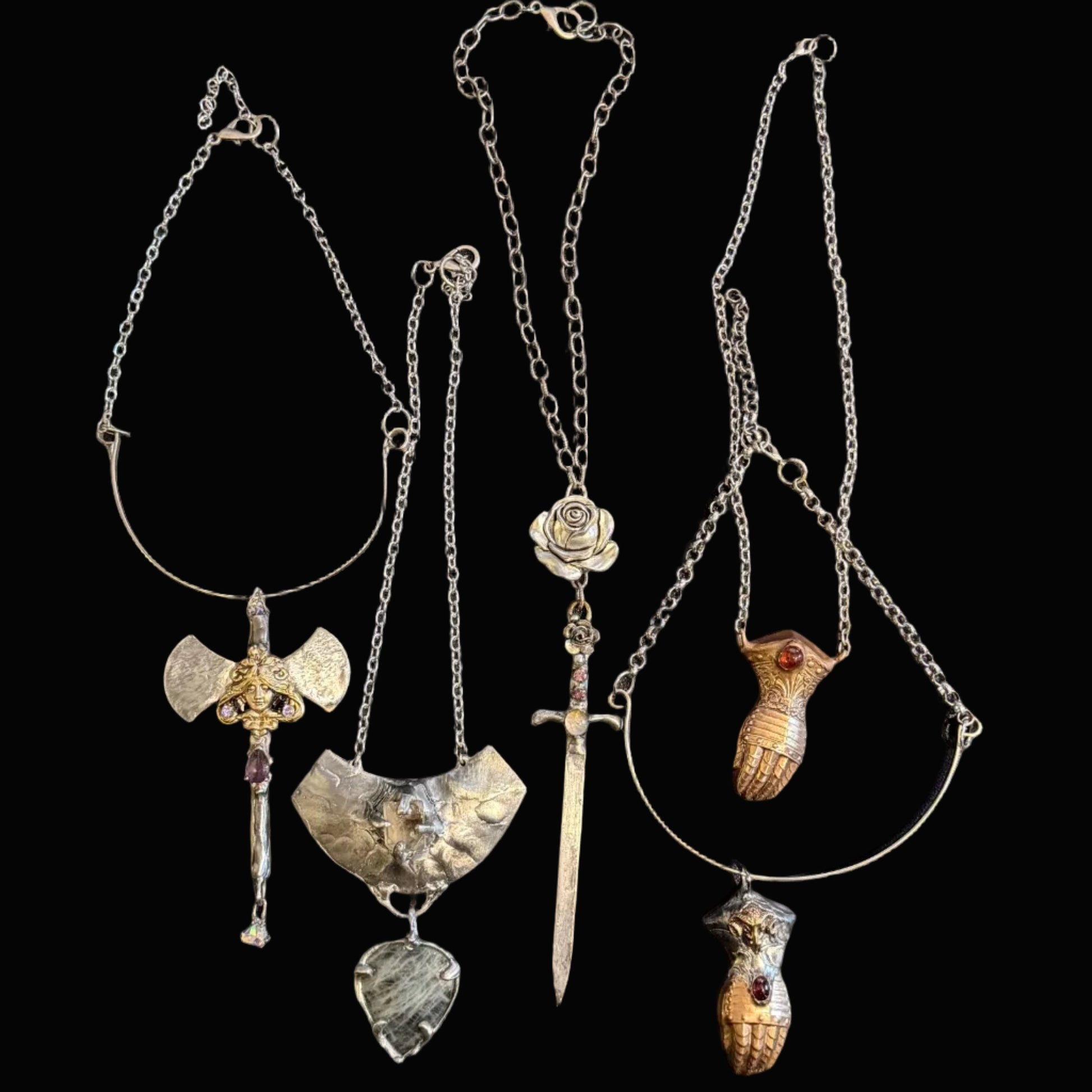 Four distinct silver-toned pendants on chains laid out on a dark desk surface, with a keyboard at the top and dark clothing at the bottom. The pendants include a winged shield, a sword, a rose, a bird, and a small animal figure.