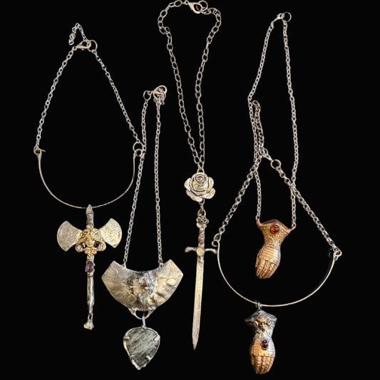 Four distinct silver-toned pendants on chains laid out on a dark desk surface, with a keyboard at the top and dark clothing at the bottom. The pendants include a winged shield, a sword, a rose, a bird, and a small animal figure.