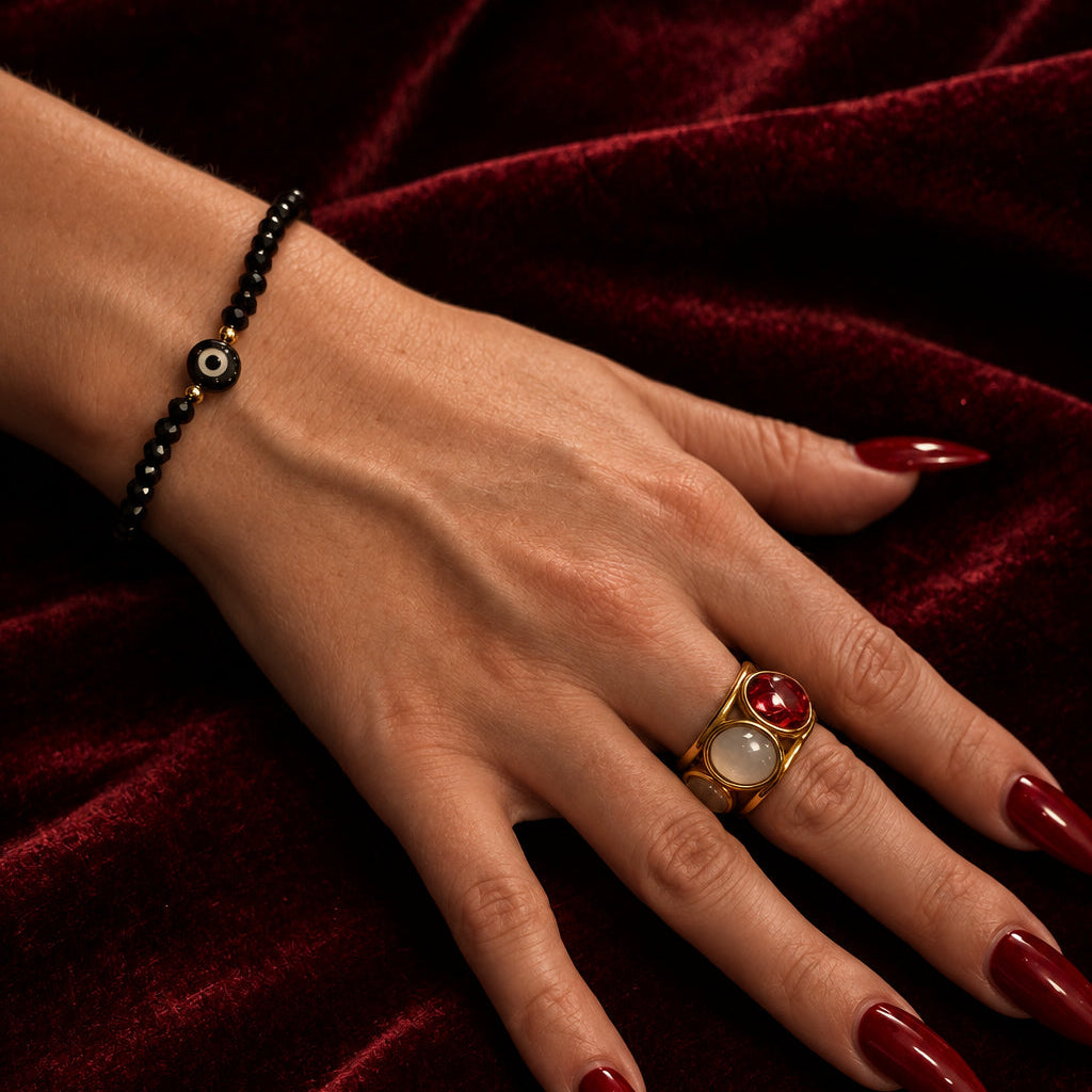 A close-up of a hand with long red manicured nails resting on a dark brown wooden surface, wearing a gold signet ring with a red heart and white oval center, and a black beaded bracelet with a circular charm.
