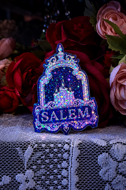 A holographic enamel pin shaped like an ornate archway with a castle inside and the word 'SALEM' at the bottom, displayed on a lace surface in front of red and pink roses.
