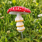 A decorative mushroom-shaped metal pin with a red cap, white spots, and a beige-brown stem, photographed on a textured brown surface.