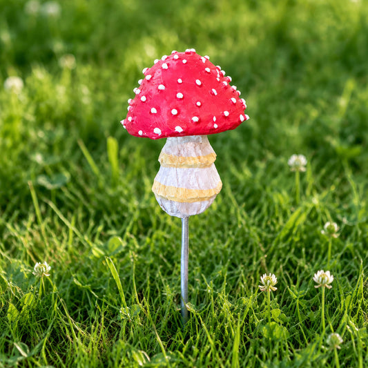 A mushroom-shaped decorative object with a red cap covered in white polka dots, a textured beige and yellow striped stem, and a silver metal rod extending from the side, placed on a brown textured surface.