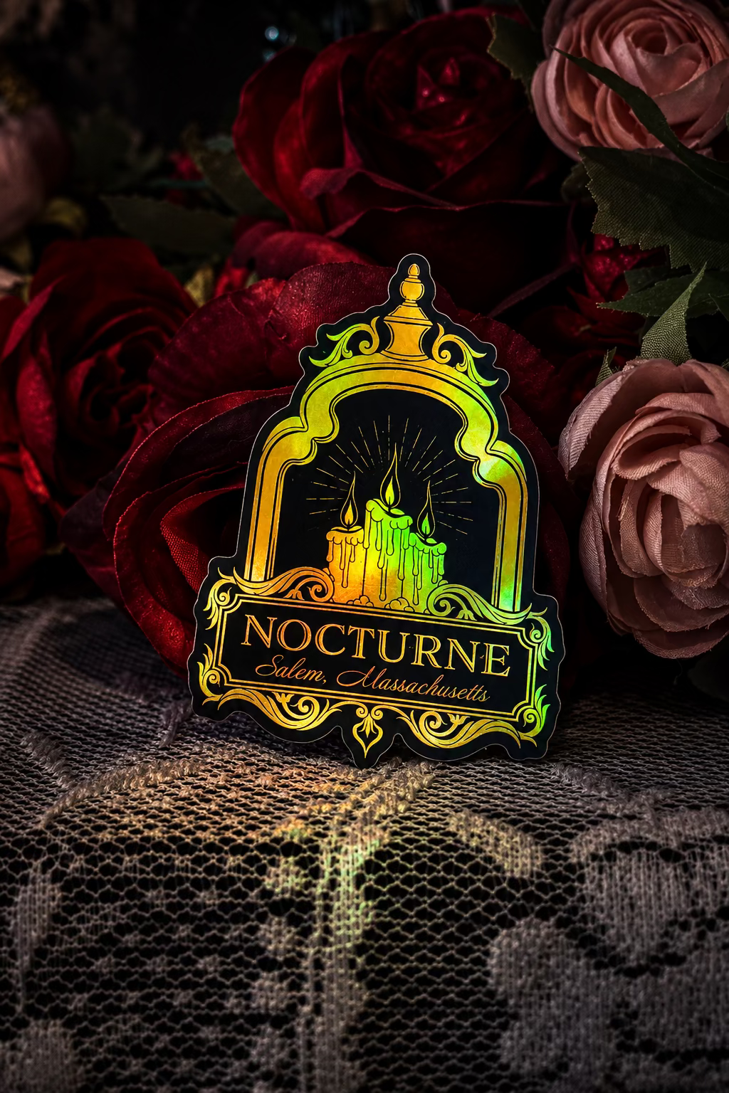 A die-cut sticker or pin featuring a castle under an ornate arch with the text 'NOCTURNE Salem, Massachusetts' in gold on a dark background, styled on white lace fabric with red and pink roses behind it.
