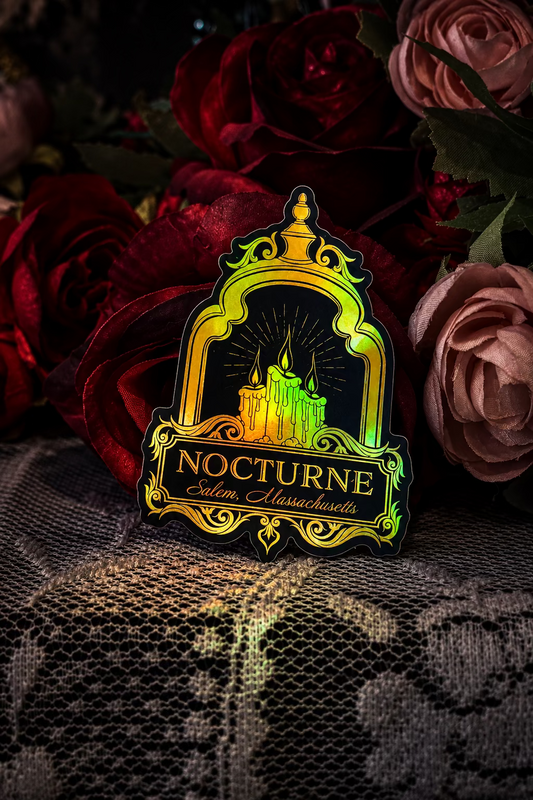 A die-cut sticker or pin featuring a castle under an ornate arch with the text 'NOCTURNE Salem, Massachusetts' in gold on a dark background, styled on white lace fabric with red and pink roses behind it.