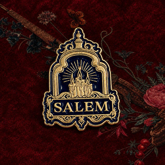 A decorative enamel pin shaped like a Salem cottage under a decorative arch, with the word 'SALEM' at the bottom. The pin is dark blue and gold-toned, photographed on a light textured background with dried botanical elements and a butterfly illustration.