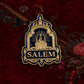 A decorative enamel pin shaped like a Salem cottage under a decorative arch, with the word 'SALEM' at the bottom. The pin is dark blue and gold-toned, photographed on a light textured background with dried botanical elements and a butterfly illustration.