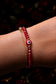 Close-up of a wrist wearing a red beaded bracelet with gold chain, against a blurred fruit background.